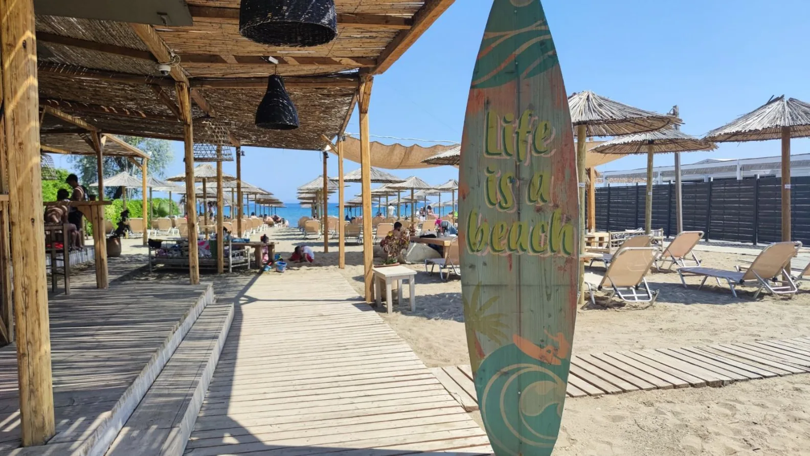 Holzsteg einer Beach-Bar in Schinias mit Strohschirmen, Liegen im Sand und dem Meer im Hintergrund, daneben ein Surfbrett mit der Aufschrift Life is a beach