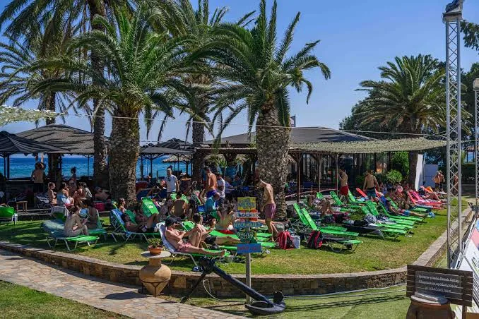 Die Beach-Bar Karavi in Schinias: grüne Liegen auf Rasen unter Palmen, viele Gäste, das hölzerne Boot als Wahrzeichen und das Meer im Hintergrund