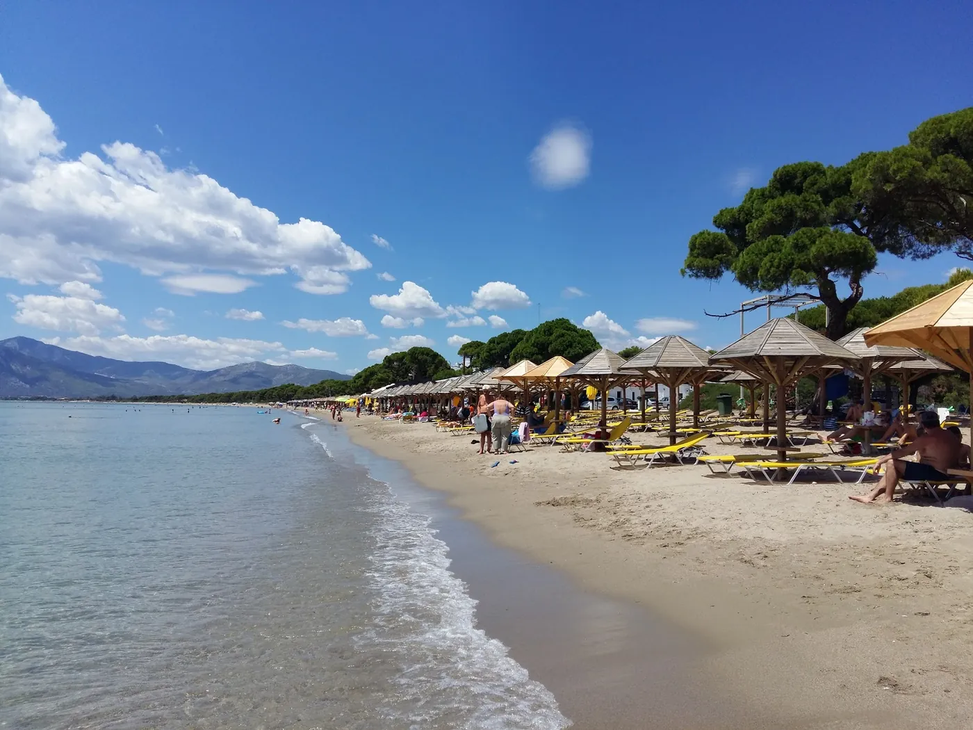 Der lange Sandstrand von Schinias: flaches türkisgrünes Wasser, Strohschirme mit gelben Liegen, Pinien und Berge im Hintergrund