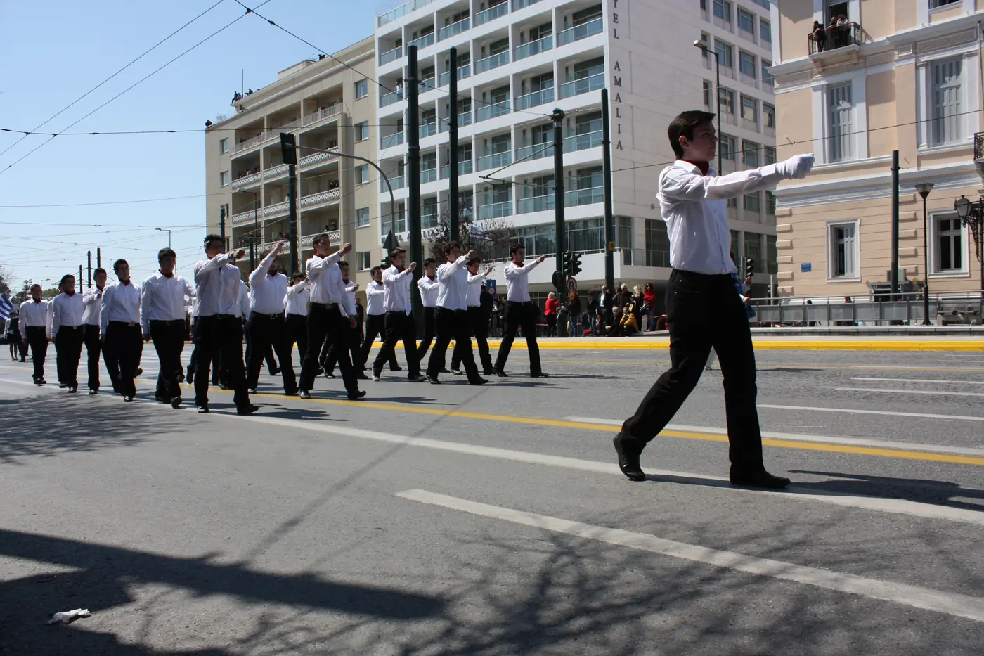 Μαθήτριες με λευκές μπλούζες και μπλε φούστες παρελαύνουν σε φαρδύ δρόμο της Αθήνας με πολυκατοικίες