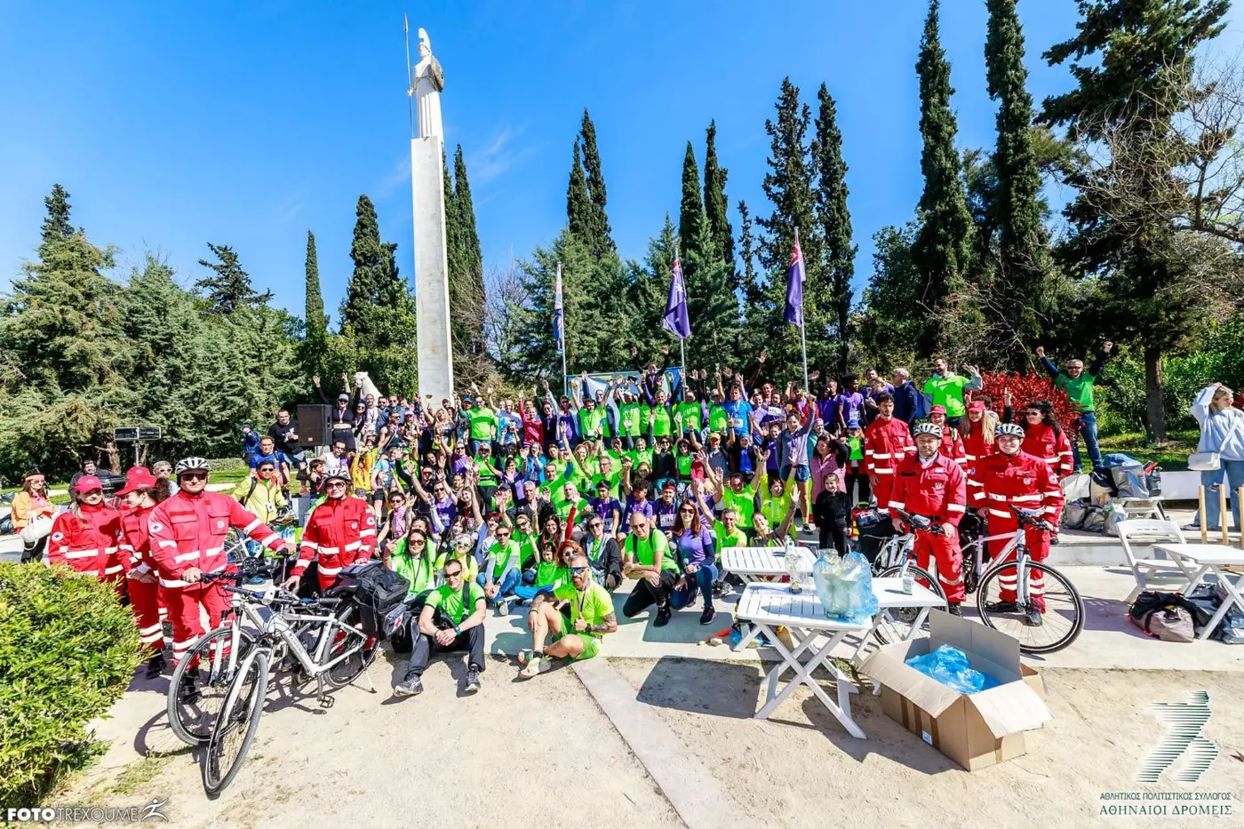 Corredores de t-shirts verdes e roxas, voluntários da Cruz Vermelha com bicicletas e a estátua de Atena no Pedion tou Areos