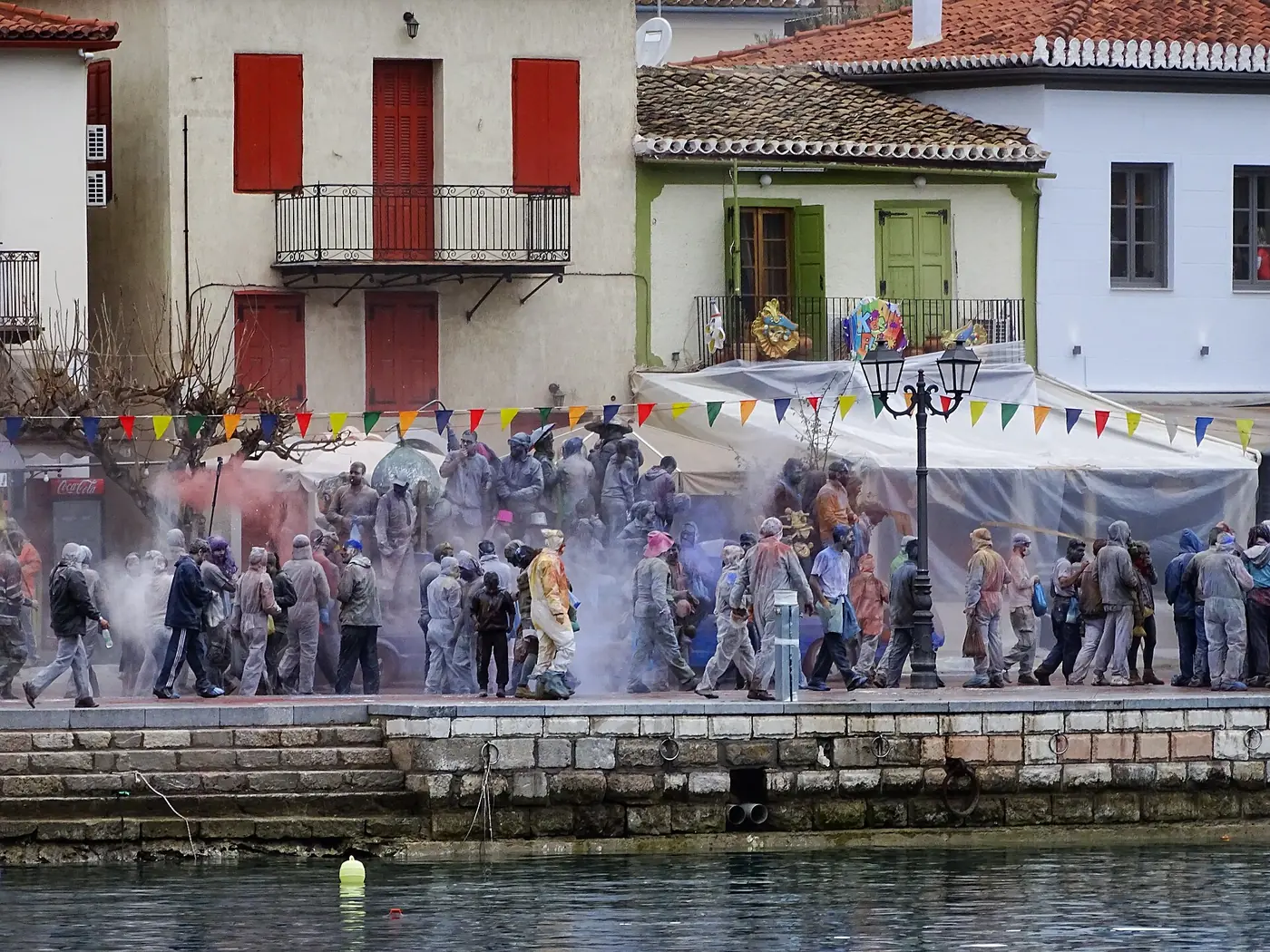 Gens couverts de farine sur le quai de Galaxidi devant des maisons néoclassiques avec des fanions colorés
