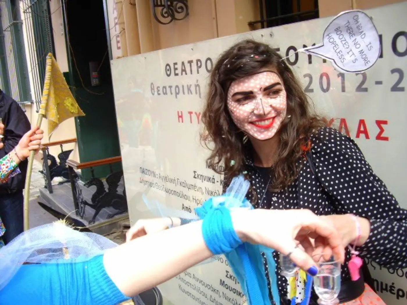 Jeune femme au visage maquillé et ruban bleu au bras souriant devant une affiche de théâtre