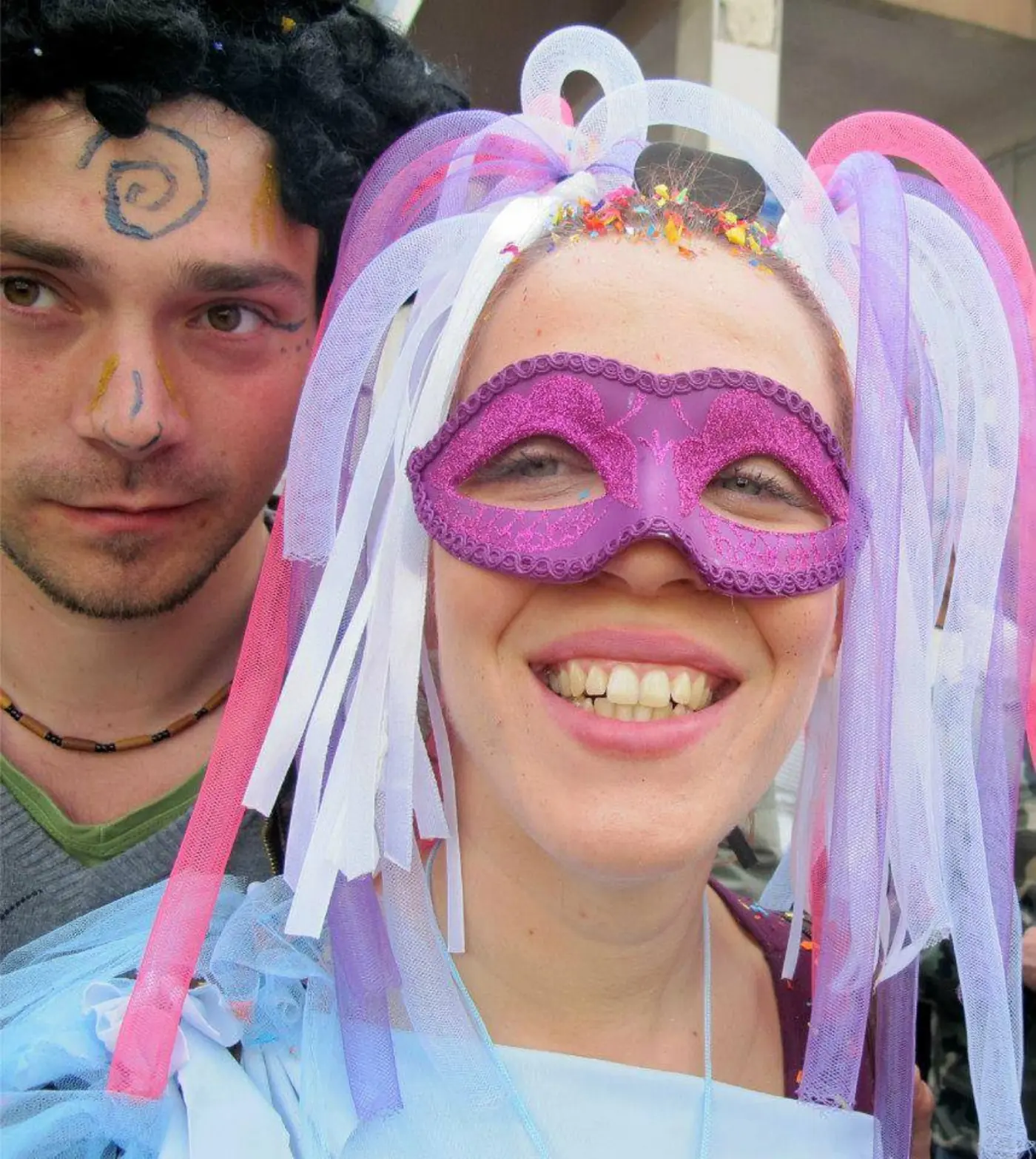 Femme souriante avec un masque violet et des rubans roses dans les cheveux lors d’un carnaval à Athènes