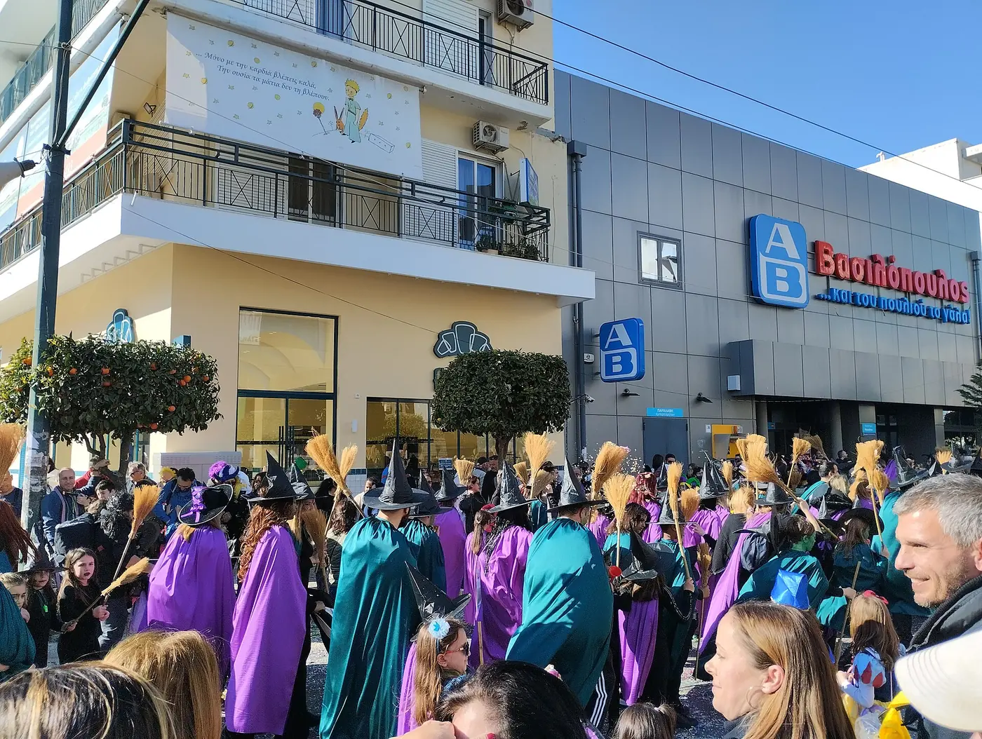Groupe d’enfants déguisés en sorcières avec capes violettes et vertes et balais défilant dans une rue de Moschato devant un supermarché