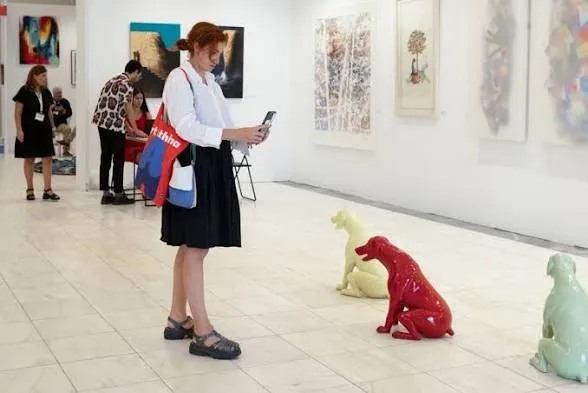 Une visiteuse photographie des œuvres au mur avec son téléphone, trois sculptures de chiens rouge, jaune et vert au sol