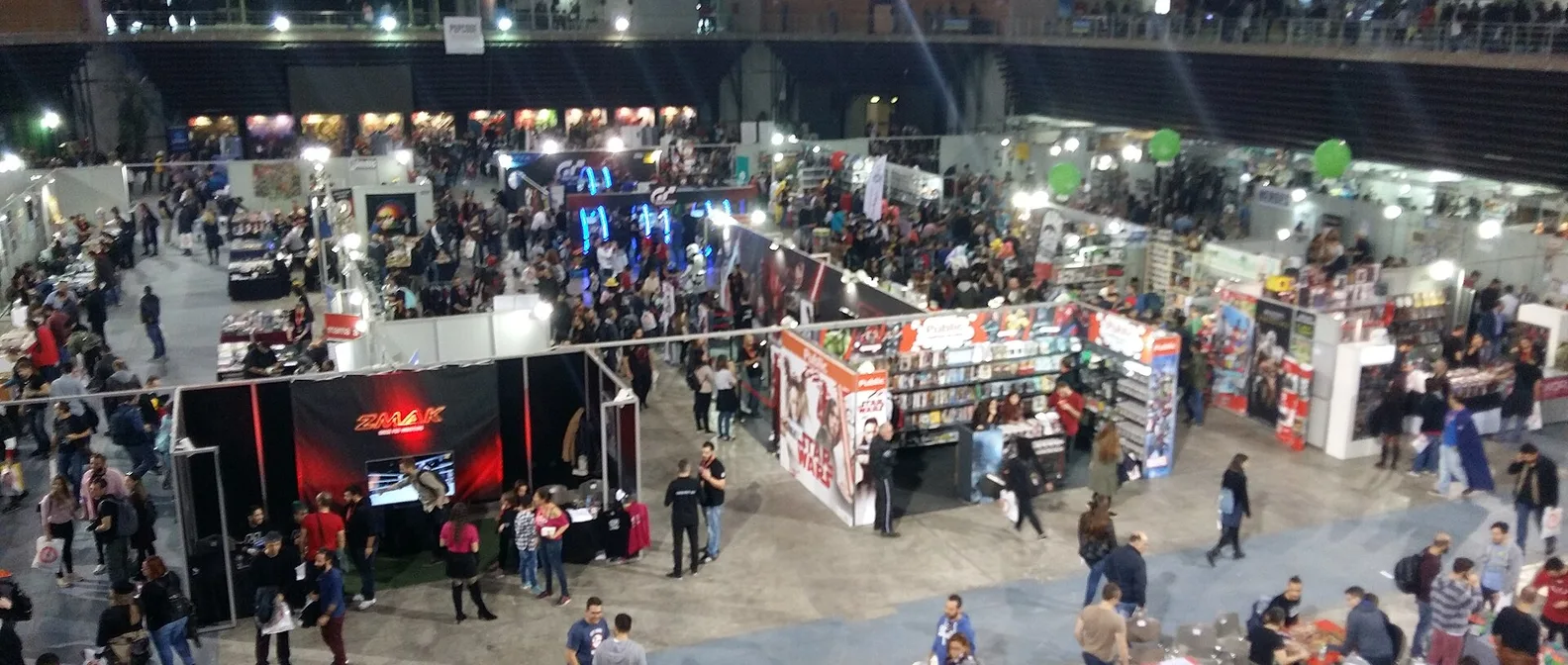 Залата на AthensCon отгоре, с щандове за комикси и игри, осветени витрини и много хора по пътеките
