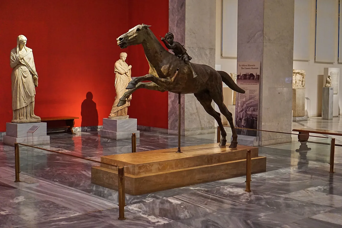Ο χάλκινος Τζόκεϊ του Αρτεμισίου, ένα άλογο σε καλπασμό με μικρό αναβάτη, σε αίθουσα με κόκκινο τοίχο και μαρμάρινα αγάλματα