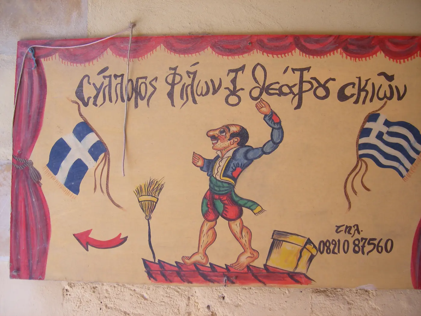 Ζωγραφισμένη αφίσα θεάτρου σκιών με τον Καραγκιόζη ανάμεσα σε δύο ελληνικές σημαίες