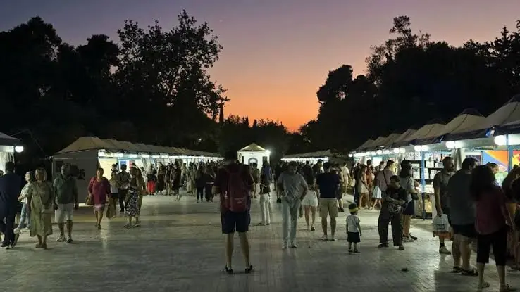 Φωτισμένα περίπτερα του Φεστιβάλ Βιβλίου στο Πεδίον του Άρεως με πορτοκαλί ουρανό στο ηλιοβασίλεμα και κόσμο στην πλατεία