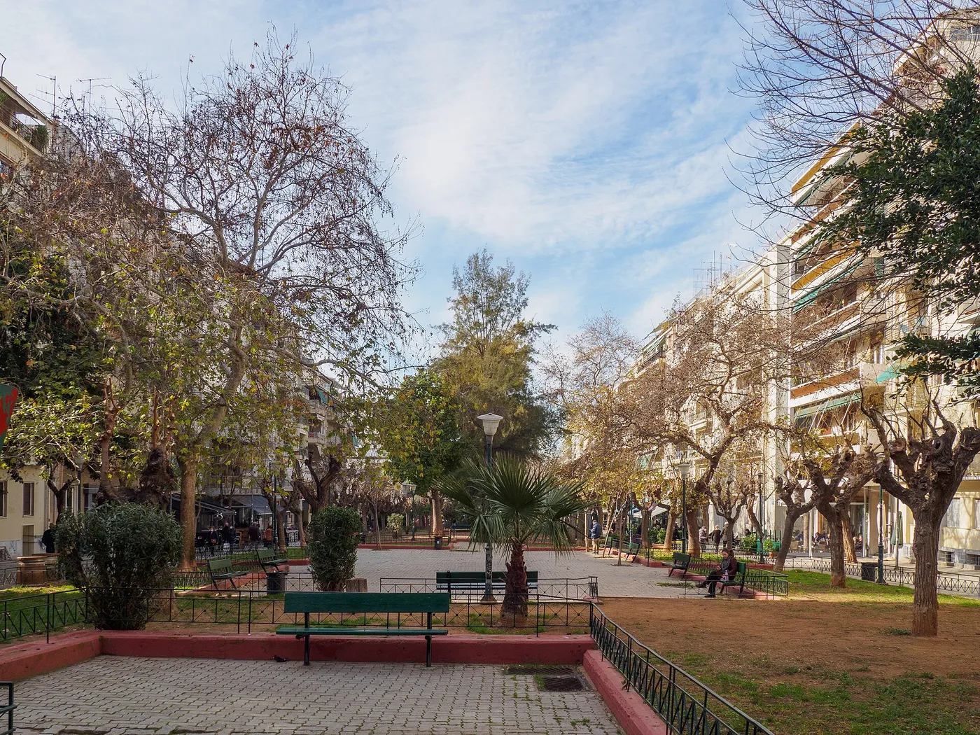 Calle peatonal con árboles, bancos y bloques de pisos en Fokionos Negri bajo un cielo azul