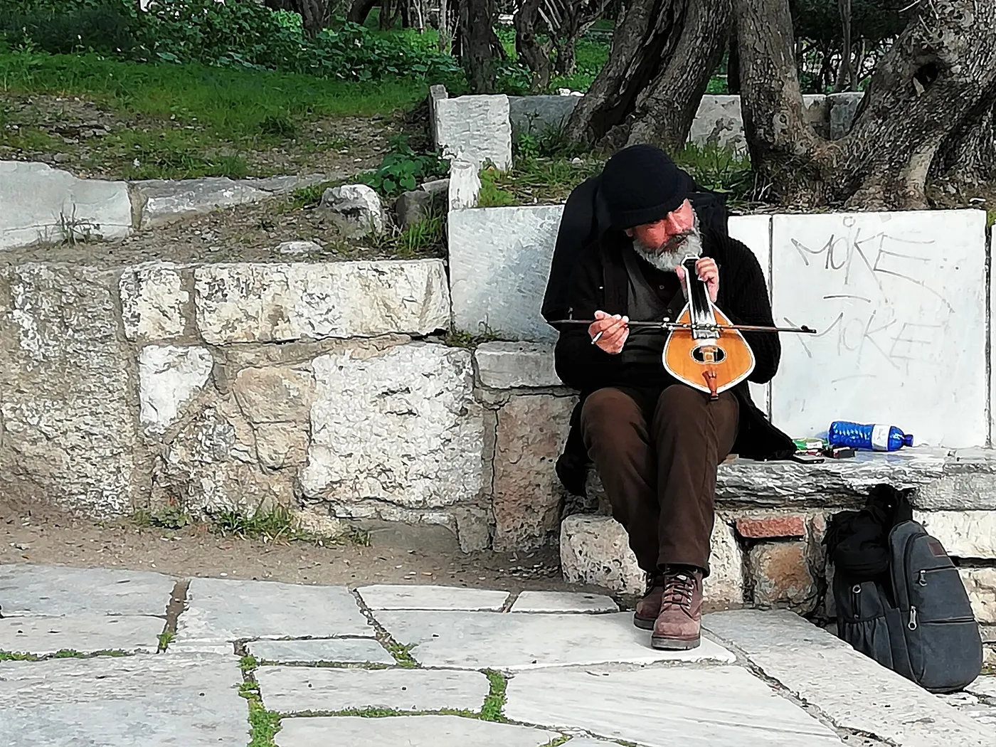 Un músico con gorro negro toca la lira sentado en un murete de mármol bajo la Acrópolis