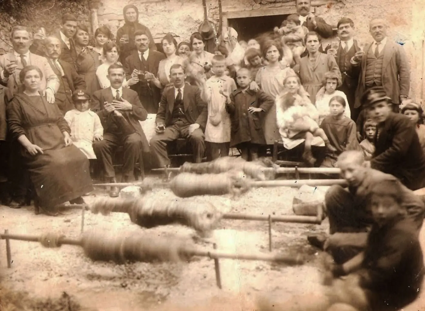 Ancienne photo sépia d’une grande famille devant une maison en pierre et de garçons qui tournent des agneaux à la broche au sol