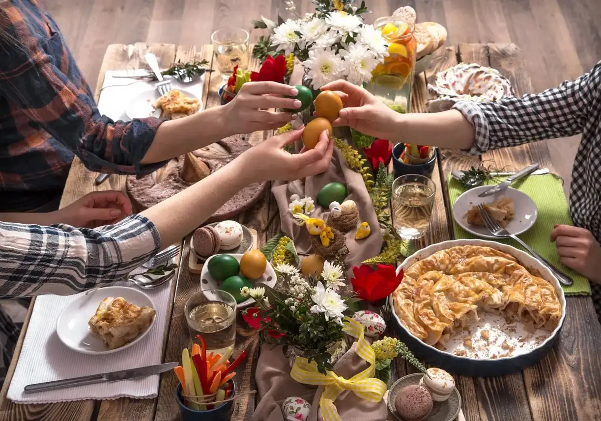Des mains entrechoquent des œufs teints au-dessus d’une table de Pâques dressée avec une tourte, des fleurs et des œufs colorés
