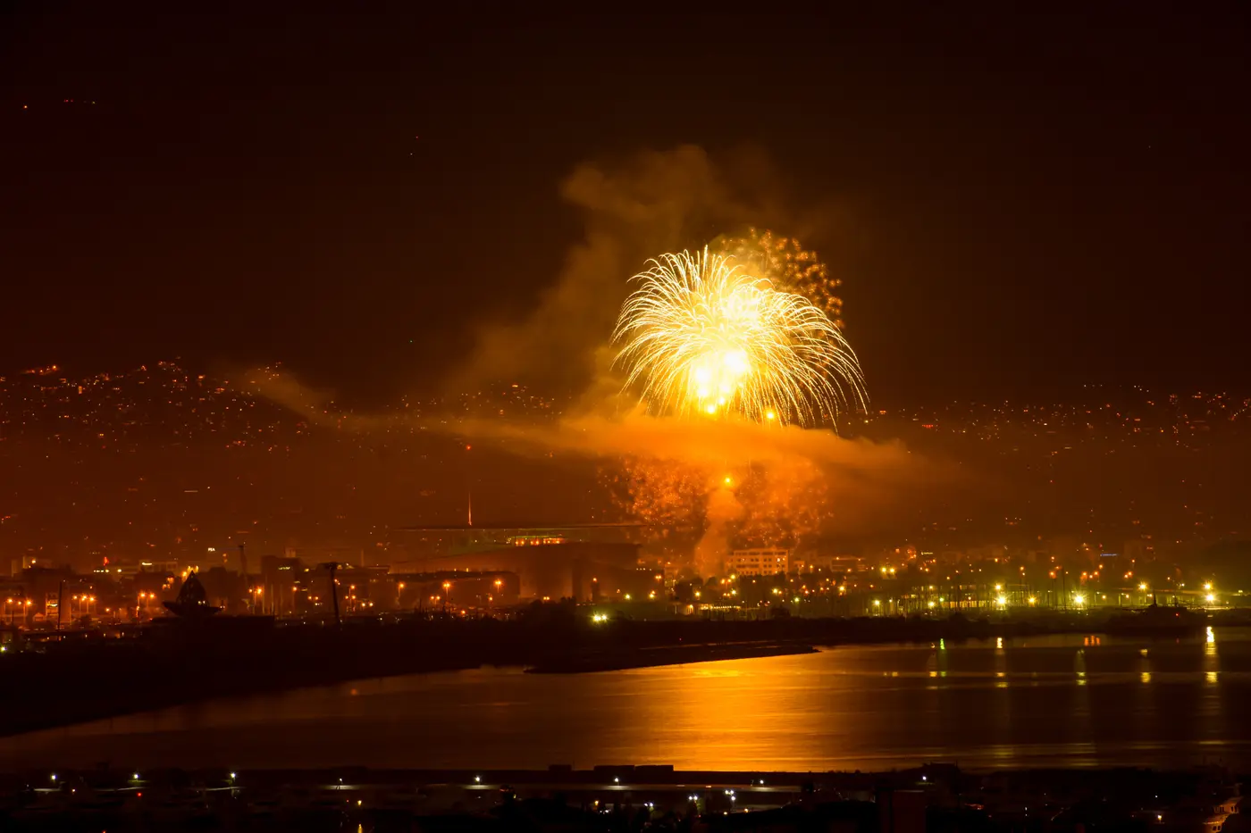 Feuerwerk über dem SNFCC in der Neujahrsnacht, die Lichter der Küste von Faliro spiegeln sich im Wasser