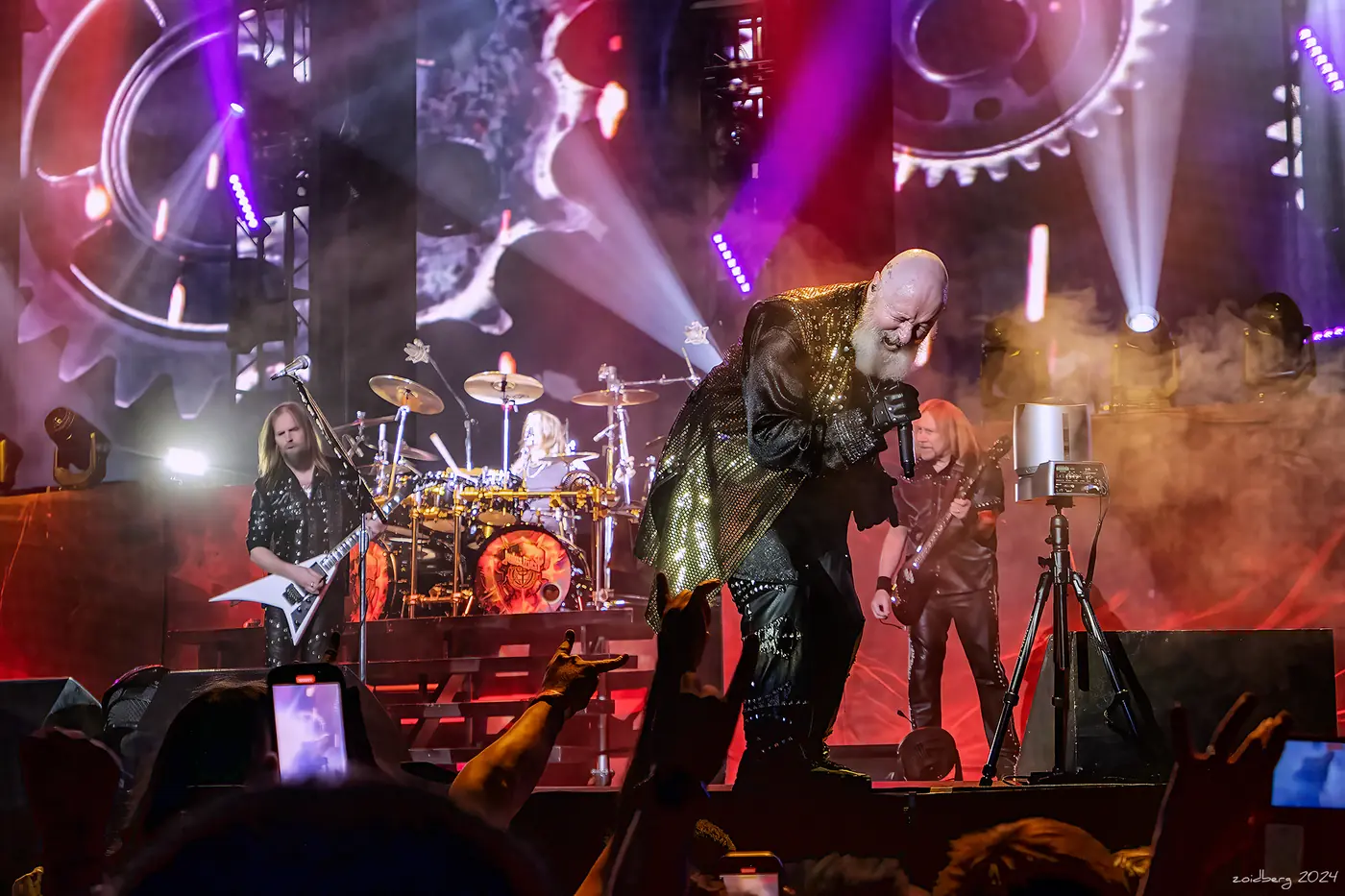 Judas Priest en el escenario bajo luces moradas y rosas, el cantante con un abrigo plateado delante de la batería