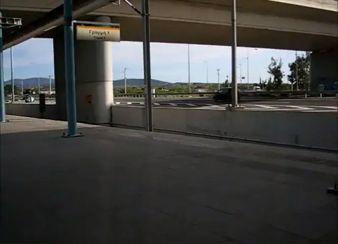 El andén vacío de la estación de Koropi con sus columnas y el cartel de la estación, con montañas al fondo