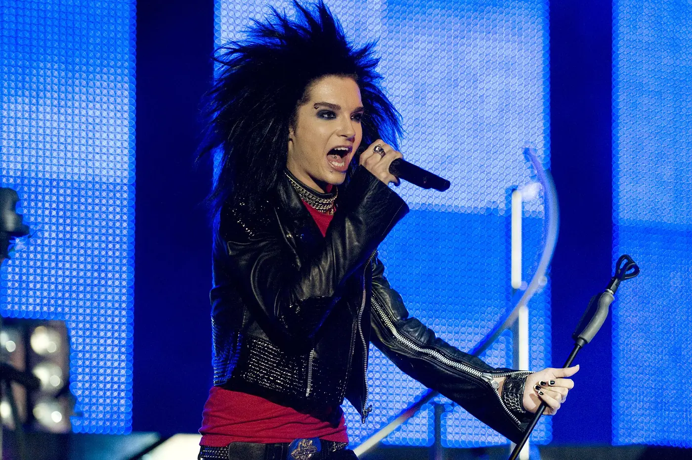 Bill Kaulitz de Tokio Hotel con chaqueta de cuero negra canta delante de una pantalla iluminada en azul