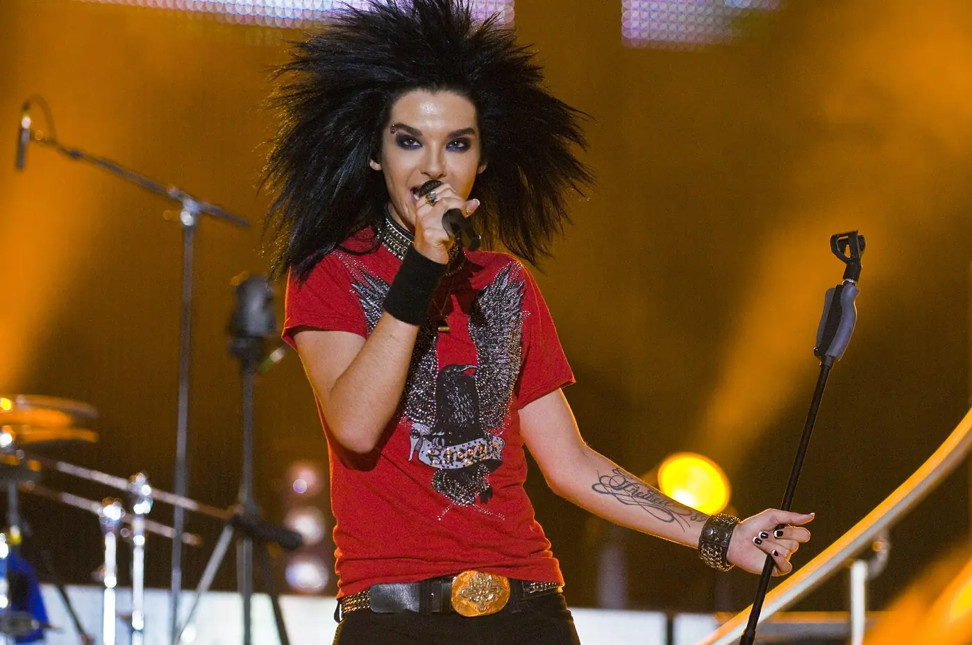 El cantante de Tokio Hotel con camiseta roja y el pelo en punta canta al micrófono sobre fondo amarillo