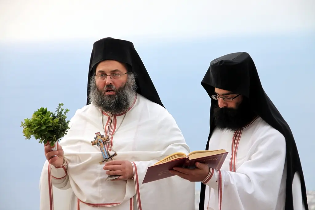 Свещеник в бели одежди държи стрък босилек и Евангелие на Великия водосвет, с морето зад него