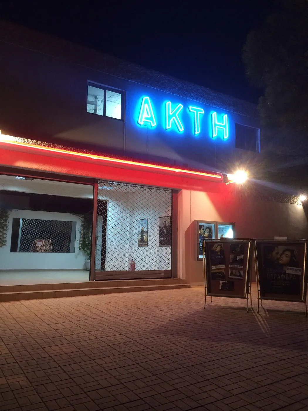 Осветеният надпис АКТИ над входа на летно кино нощем, с филмови плакати