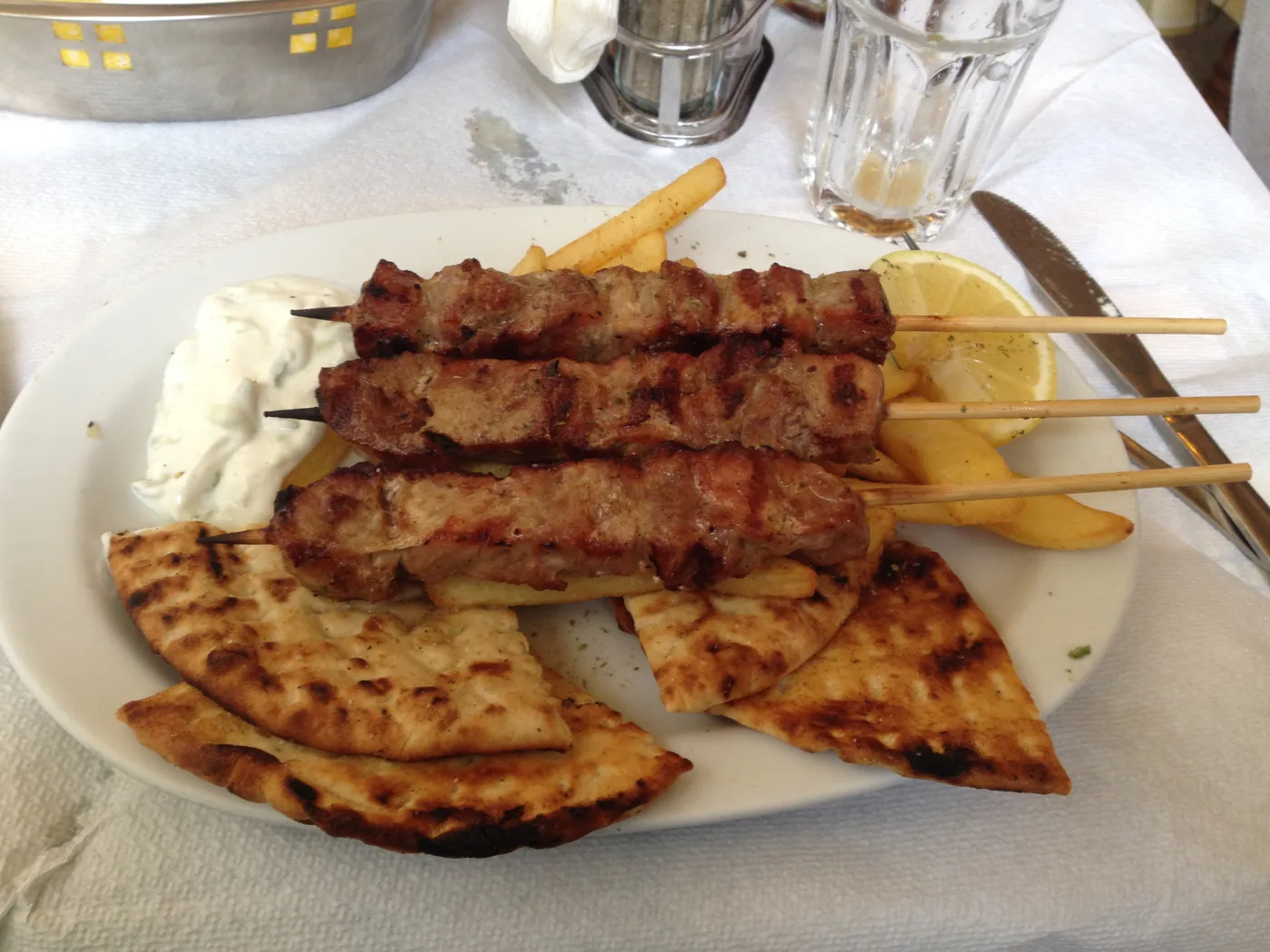 Τρία χοιρινά καλαμάκια με ψημένη πίτα, πατάτες τηγανητές, τζατζίκι και λεμόνι σε πιάτο ταβέρνας στην Αθήνα