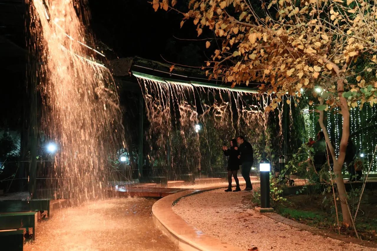 Water valt als een gordijn van een verlichte pergola in Pedion tou Areos bij nacht, met een stel dat foto’s maakt naast een vergeelde boom