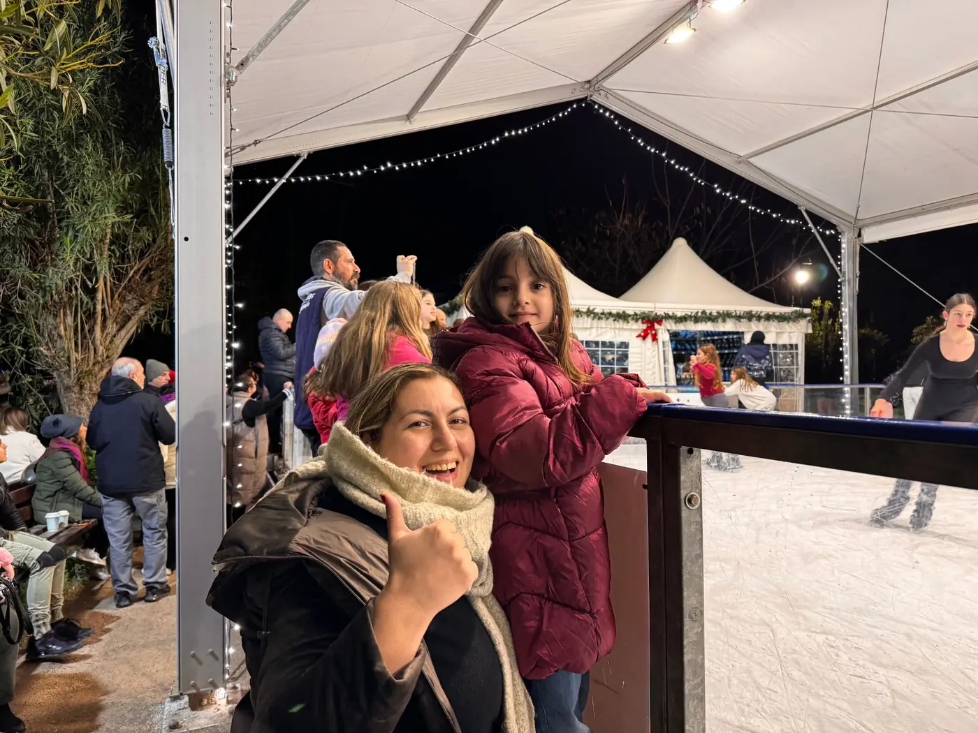 Vrouw en meisje in winterjassen bij de reling van de overdekte ijsbaan in Pedion tou Areos, met schaatsende kinderen erachter