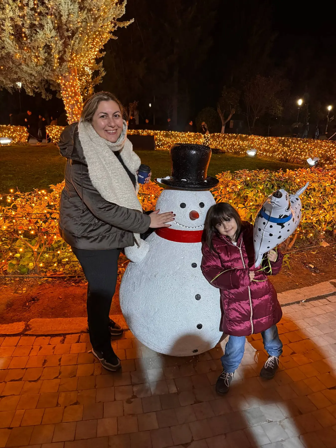 Vrouw en meisje met een ballon in de vorm van een hond poseren bij een grote sneeuwpop met zwarte hoed, voor verlichte struiken