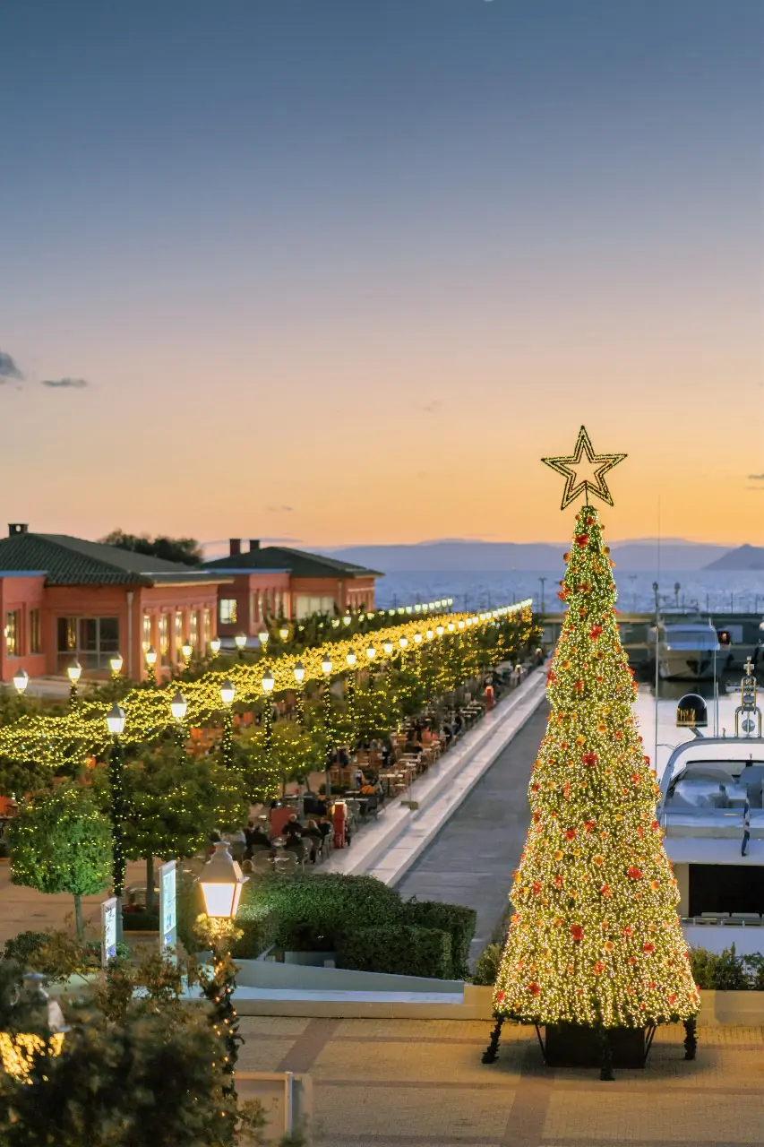 Hoge kerstboom met gouden lampjes en een ster in de top in Marina Flisvos in de schemering, met verlichte promenade, cafés en de zee op de achtergrond