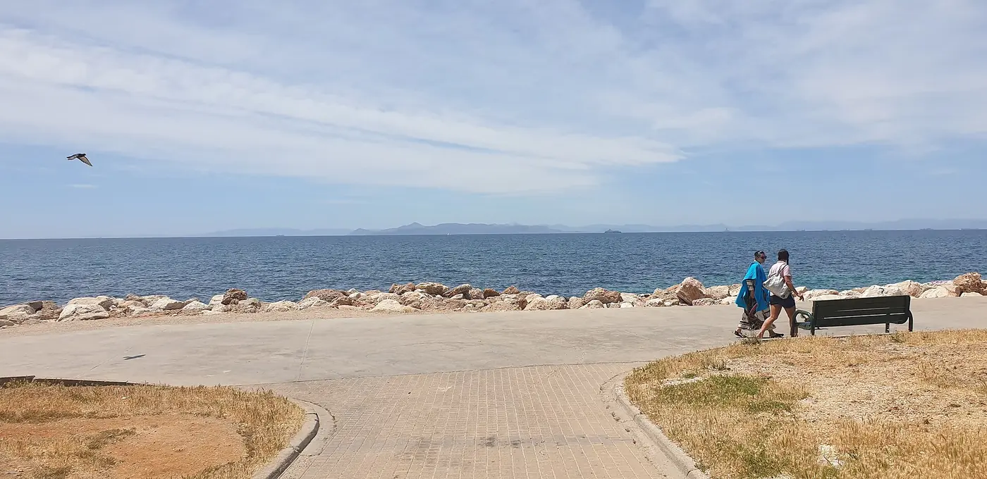 Het voetpad van het Flisvospark aan zee, met wandelaars, een bankje en de Saronische Golf op de achtergrond
