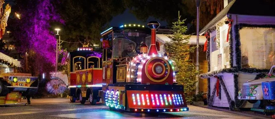 Verlicht rood treintje met gekleurde lampjes tussen versierde houten kraampjes, in het Kerstmanpark in Flisvos
