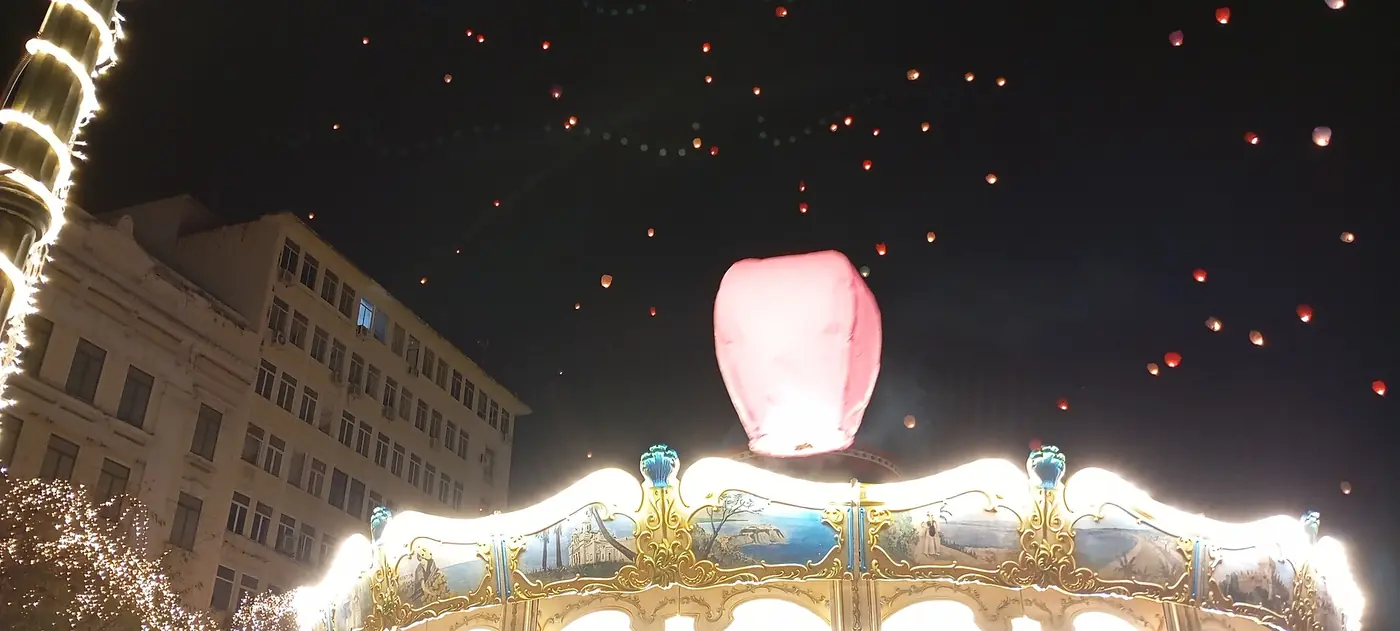 Verlichte draaimolen op het Kotziaplein bij nacht, met lantaarns in de lucht en een roze lantaarn boven het dak