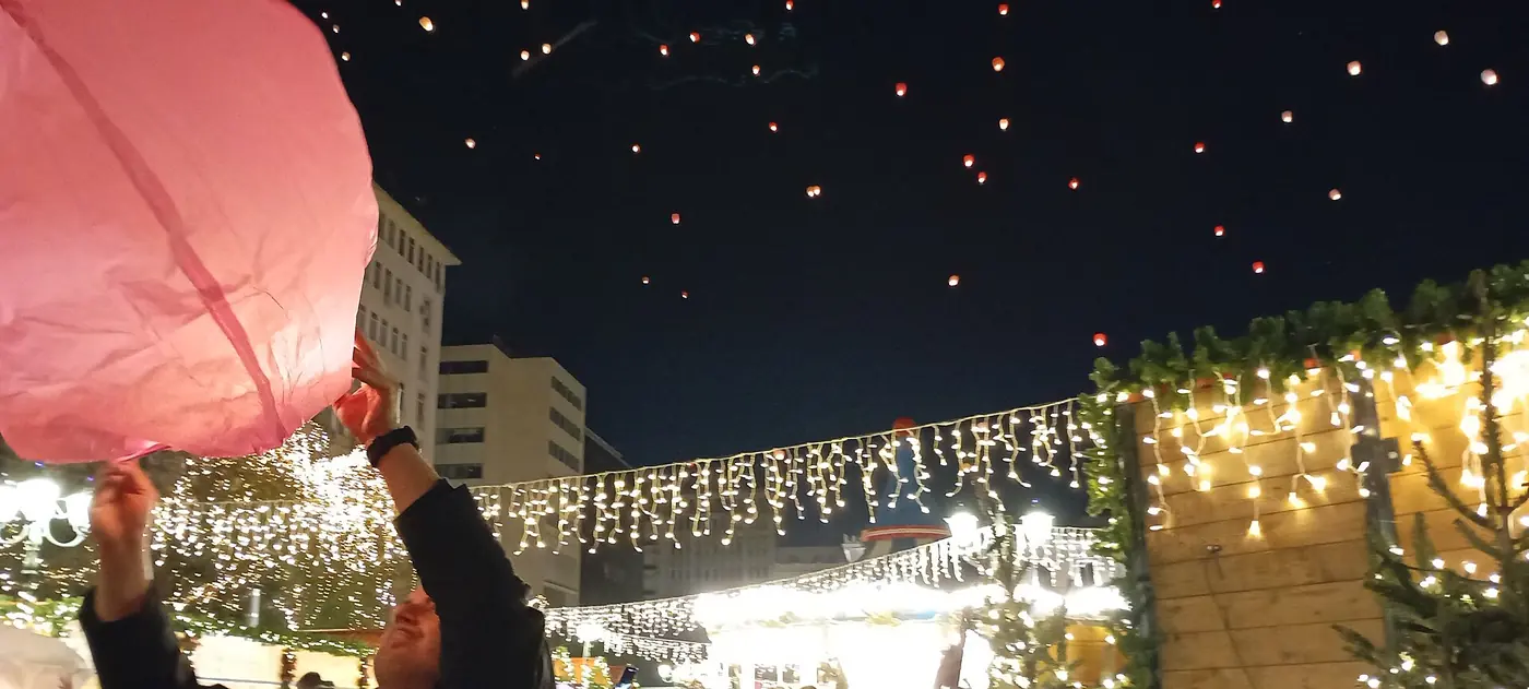 Een grote roze lantaarn in de handen van iemand, met lichtjes en houten kraampjes op de achtergrond
