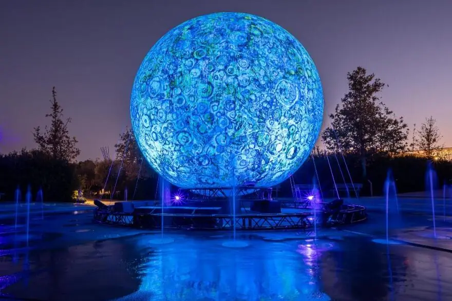 Grote blauwe lichtbol die op een maan lijkt, boven fonteinen met paarse lichten in het park van het SNFCC