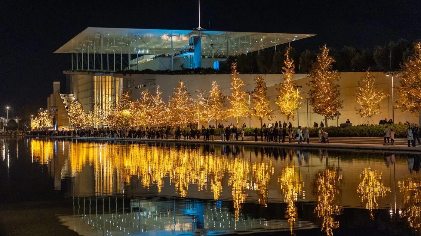 Het SNFCC-gebouw bij nacht met een rij bomen in gouden lampjes, mensen op de promenade en weerspiegelingen in het water van het Kanaal