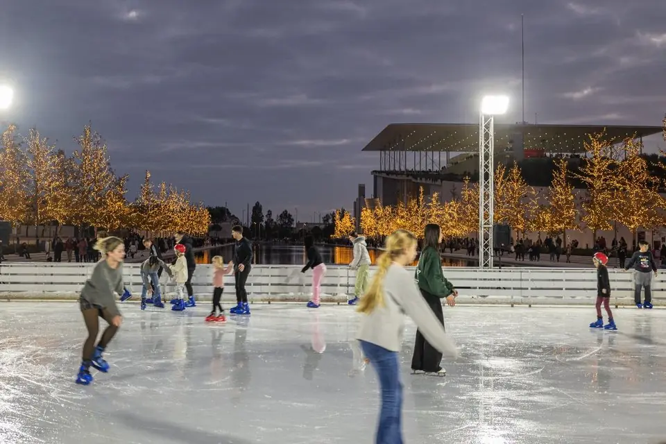 Kinderen en volwassenen schaatsen op de openluchtijsbaan van het SNFCC, met verlichte bomen en het operagebouw op de achtergrond
