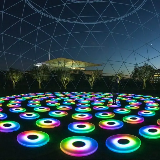 Tientallen lichtringen in alle kleuren van de regenboog op de grond, onder een doorzichtige koepel, met het SNFCC-gebouw op de achtergrond