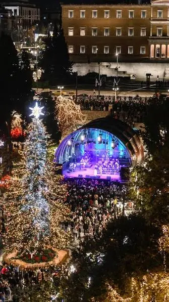 Concert op een verlicht podium op het Syntagmaplein bij nacht, met een menigte, de kerstboom links en het parlement op de achtergrond