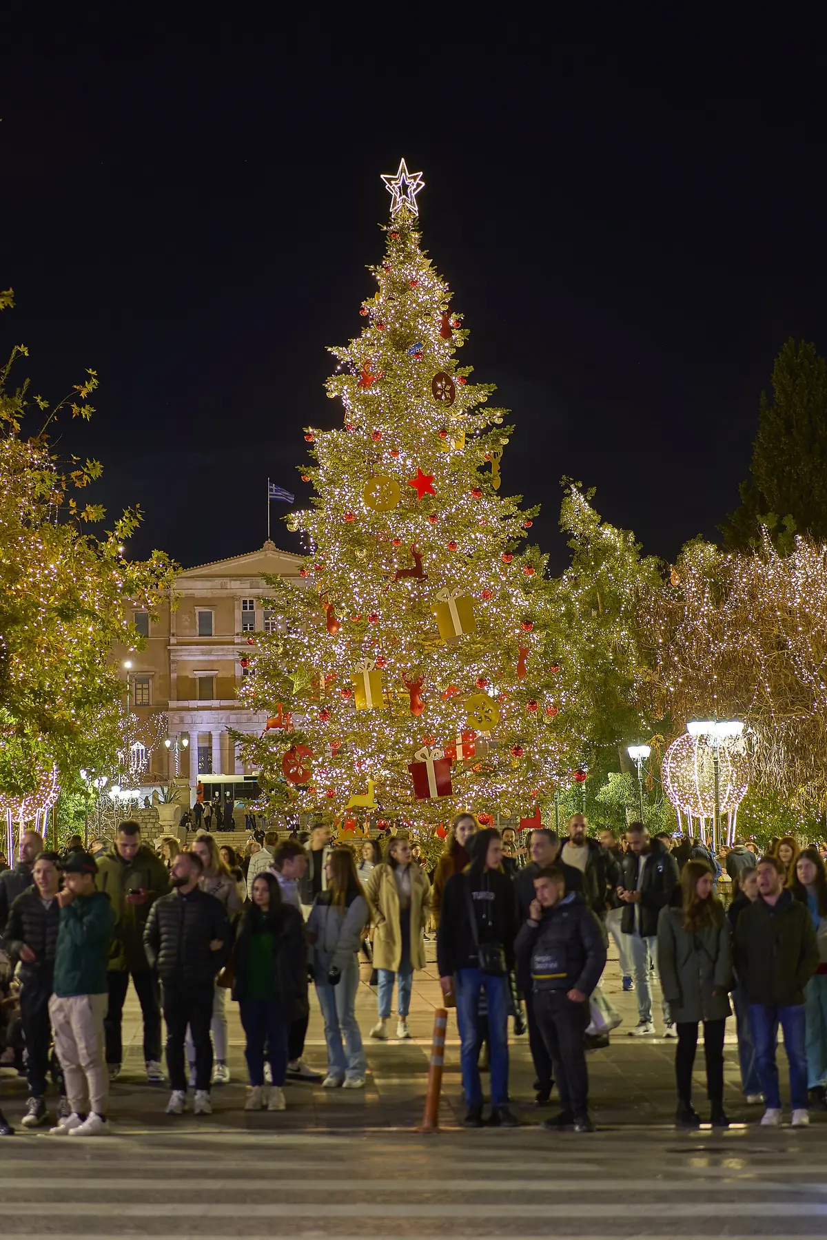 De verlichte kerstboom op het Syntagmaplein bij nacht, met mensen eromheen en het parlementsgebouw erachter