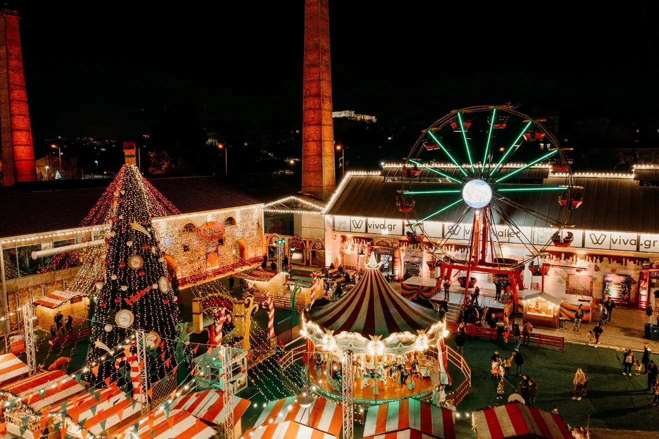 De Christmas Factory bij nacht met een boom vol klokken, een draaimolen met gestreept dak, een reuzenrad met groene lichten en de Akropolis op de achtergrond tussen de schoorstenen