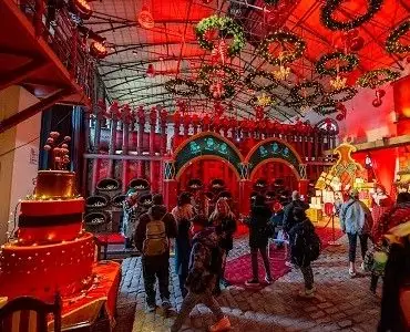 Bezoekers lopen door een versierde zaal van de Christmas Factory met rood licht, kransen aan het plafond en een grote nep-taart