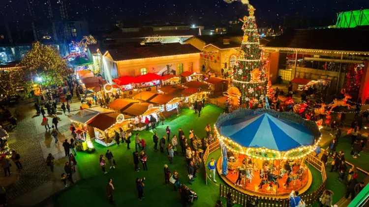Het kerstdorp van Technopolis van bovenaf bij nacht, met verlichte draaimolen, versierde boom, houten kraampjes en mensen op het gras