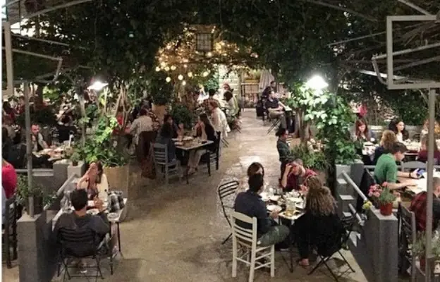Κόσμος καθισμένος το βράδυ στα τραπέζια της αυλής του Άμα Λάχει ανάμεσα σε γλάστρες και φυτά