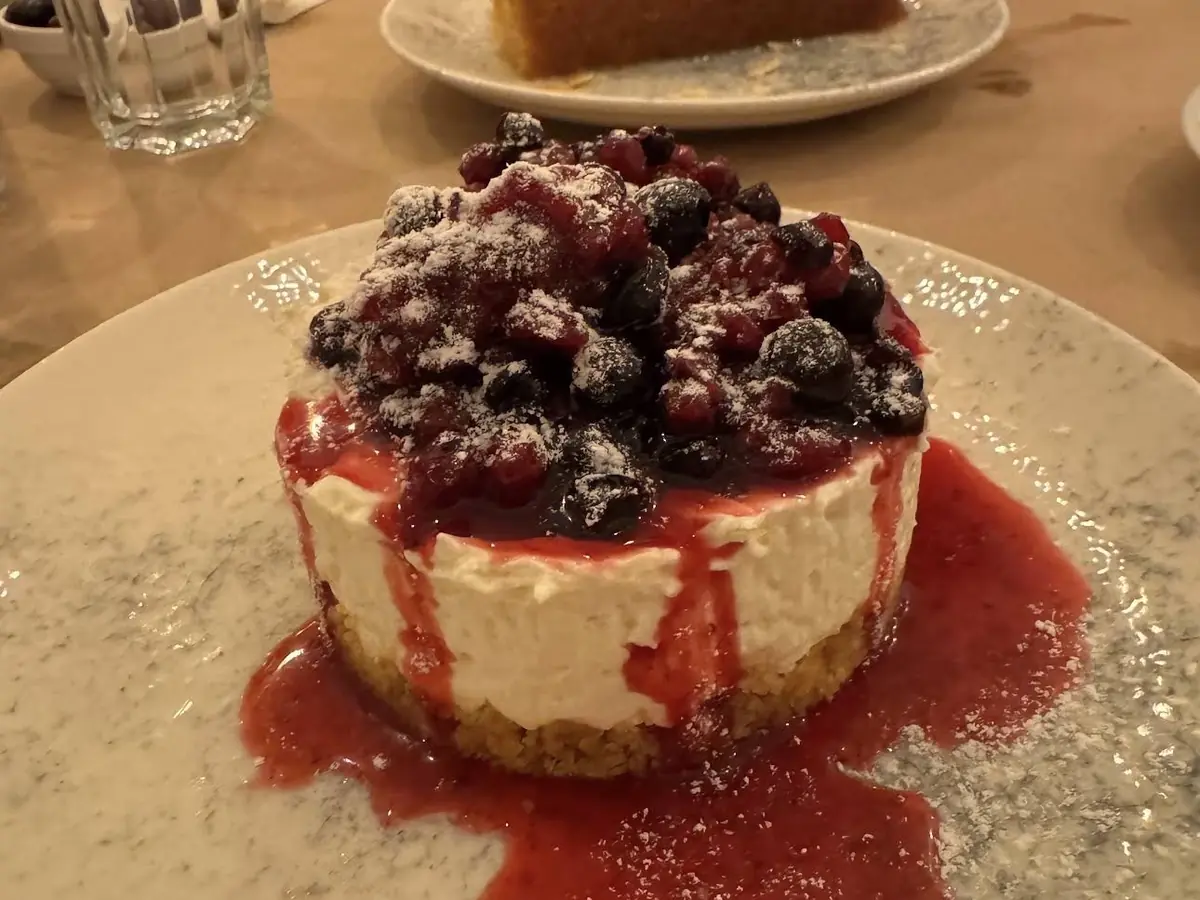 Cheesecake με φρούτα του δάσους και σιρόπι σε πιάτο, με άλλο γλυκό στο βάθος του τραπεζιού
