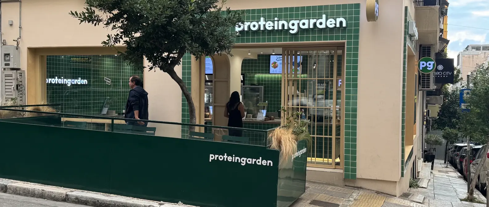 Κατάστημα Protein Garden σε γωνία με πράσινα πλακάκια, ταμπέλα proteingarden και εξωτερικό πεζούλι με φυτά