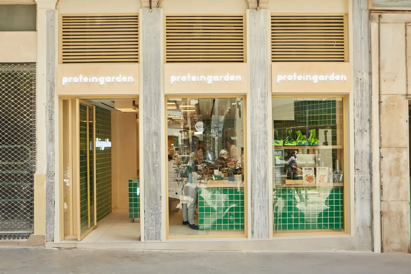 Η πρόσοψη του Protein Garden στην Κολοκοτρώνη 33, με τρία φωτεινά πινακίδια proteingarden ανάμεσα σε μαρμάρινες κολώνες