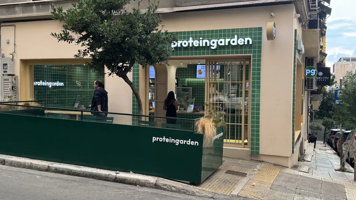 Το γωνιακό κατάστημα Protein Garden στη Σκουφά 53 στο Κολωνάκι, με πράσινα πλακάκια, ταμπέλα proteingarden και πεζούλι με φυτά