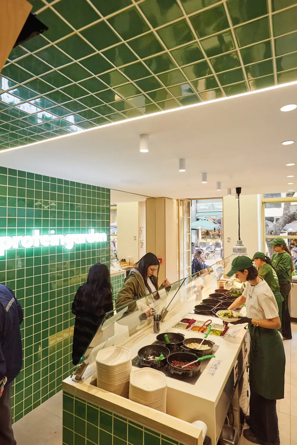 Εσωτερικό του Protein Garden στη Χέιδεν 8: πάγκος με τηγάνια και υλικά, υπάλληλοι με πράσινες ποδιές και πελάτες μπροστά στον πάγκο
