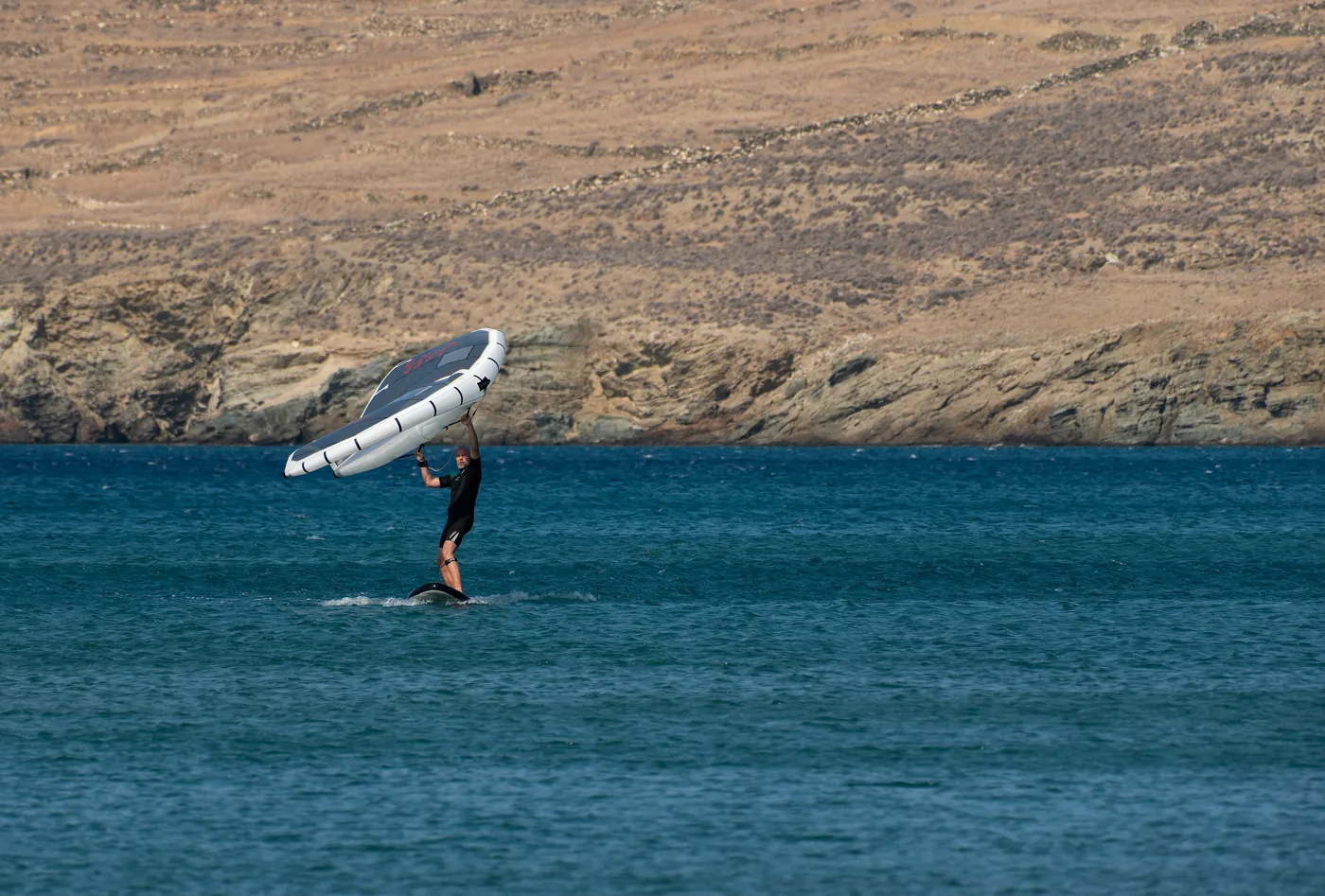 Uma pessoa a fazer wingfoil na água azul de Kionia, com colinas atrás